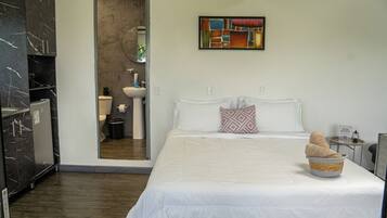 A modern bedroom with a large bed, a small bedside table, a wall-mounted painting, and a bathroom visible through an open door.