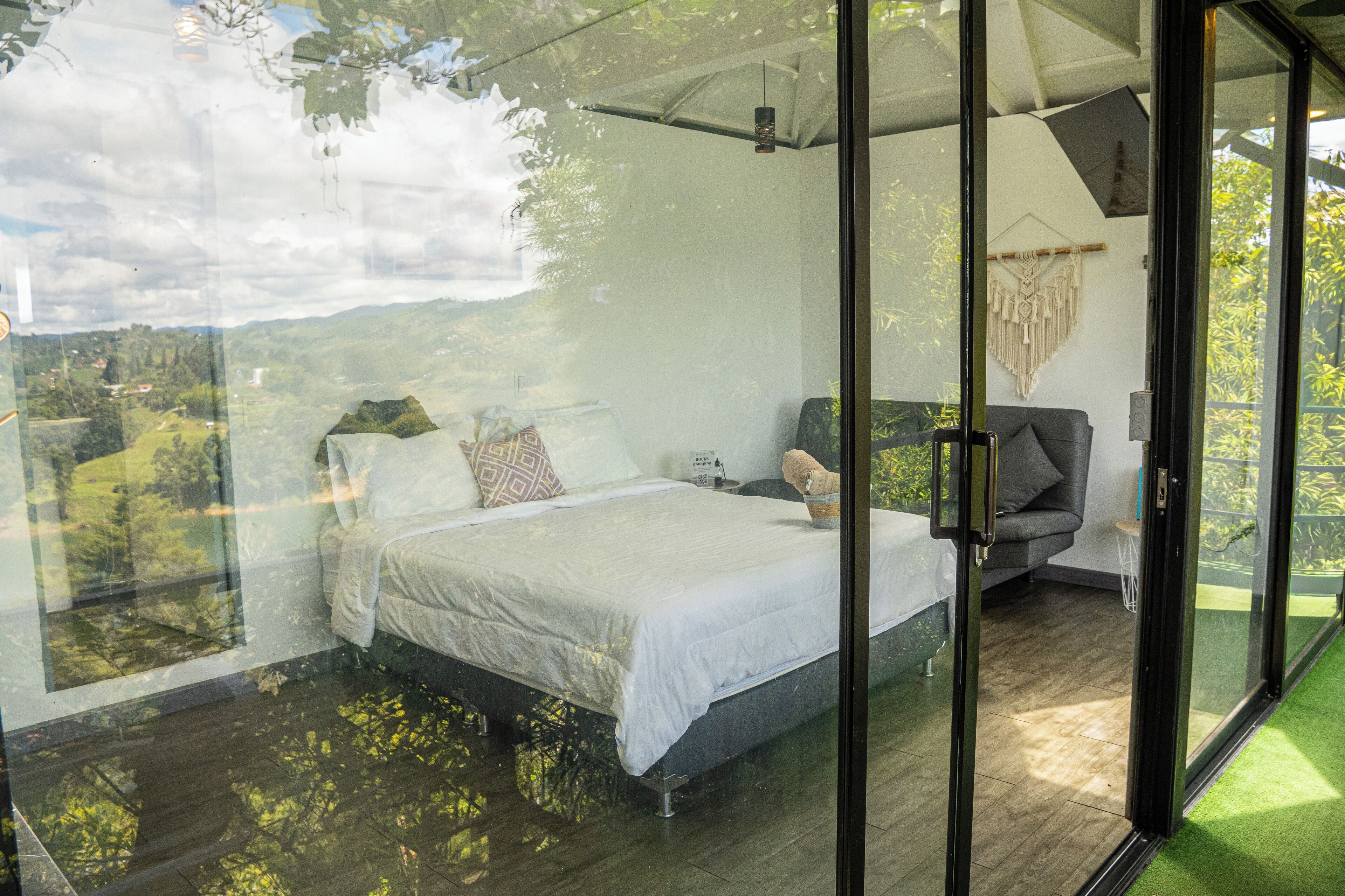 A bedroom with a bed, a nightstand, and a chair. There is a large window with a view of greenery.