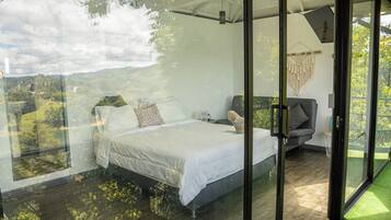 A bedroom with a bed, a nightstand, and a chair. There is a large window with a view of greenery.