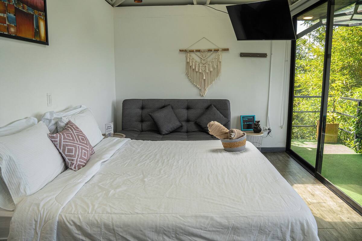 A bedroom with a large bed, grey headboard, white bedding, a macrame wall hanging, and a view of greenery through a sliding glass door.