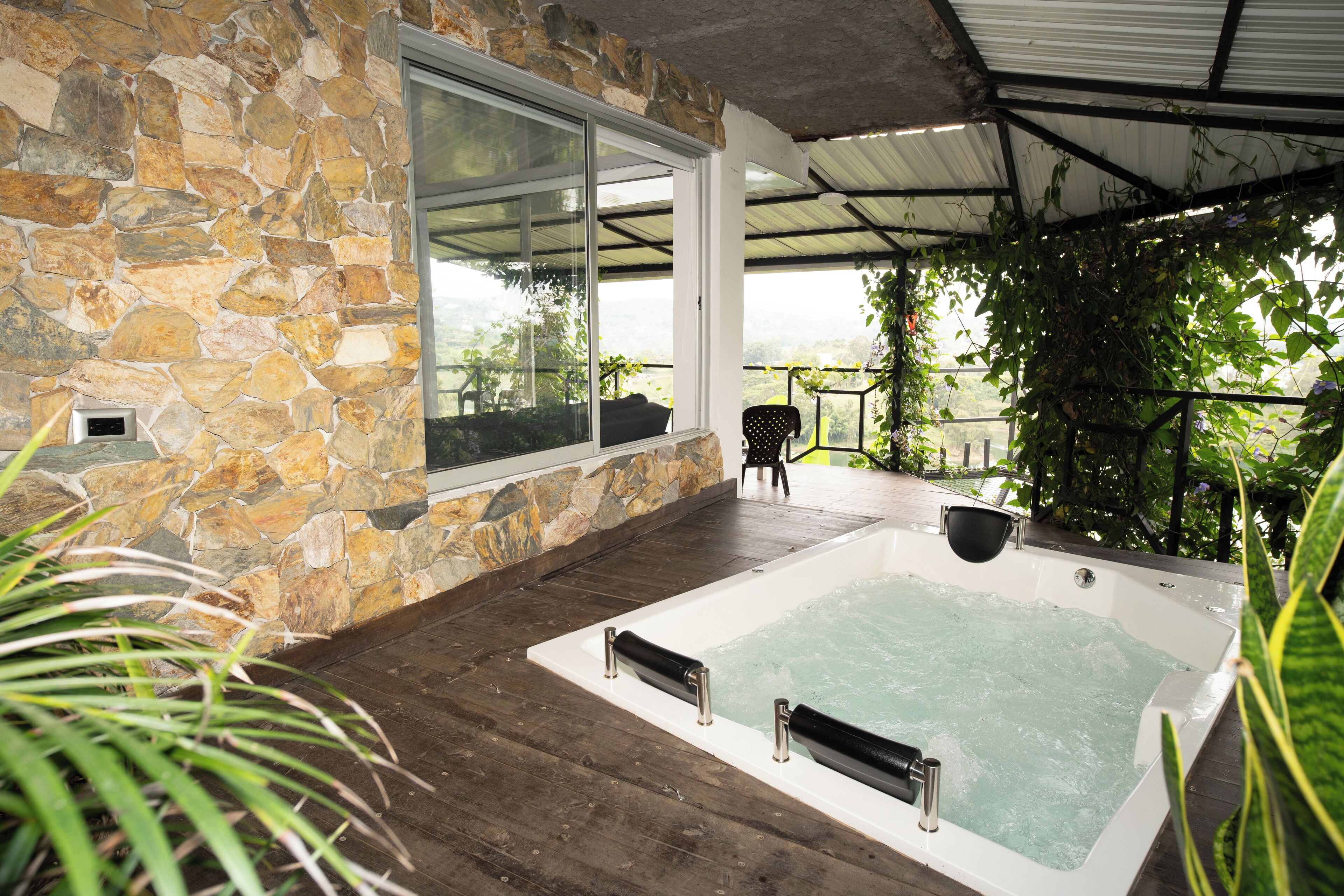 A stone-walled room with a large window, a hot tub, and a wooden deck.
