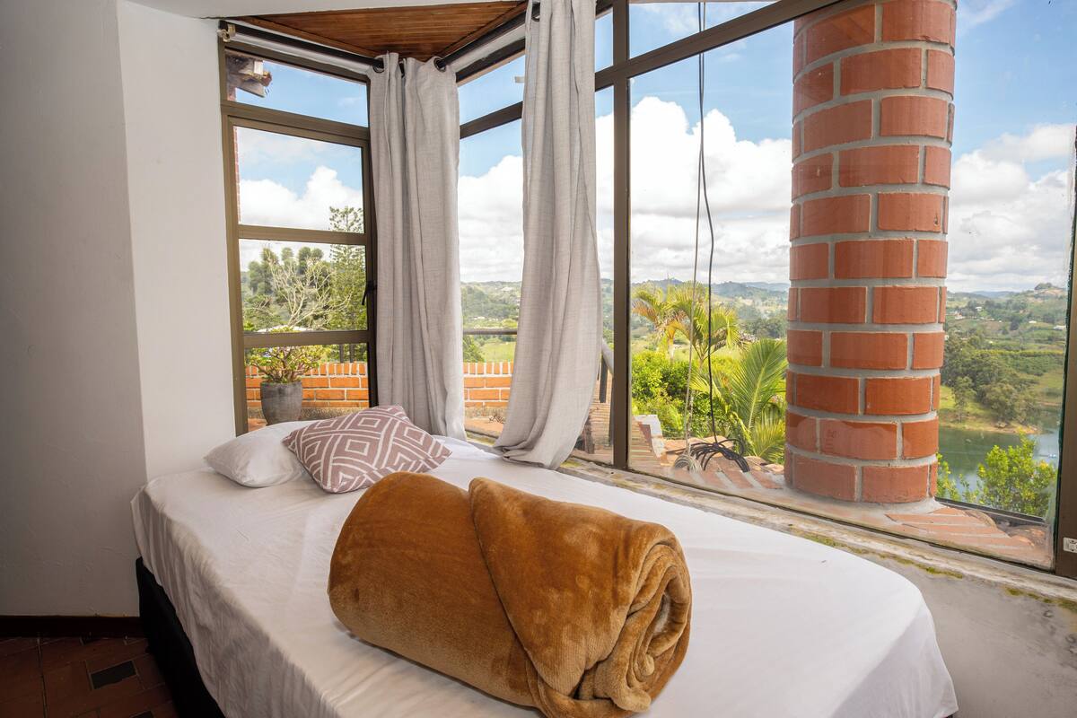 A bedroom with a large window, a bed with a brown blanket, and a view of a landscape.