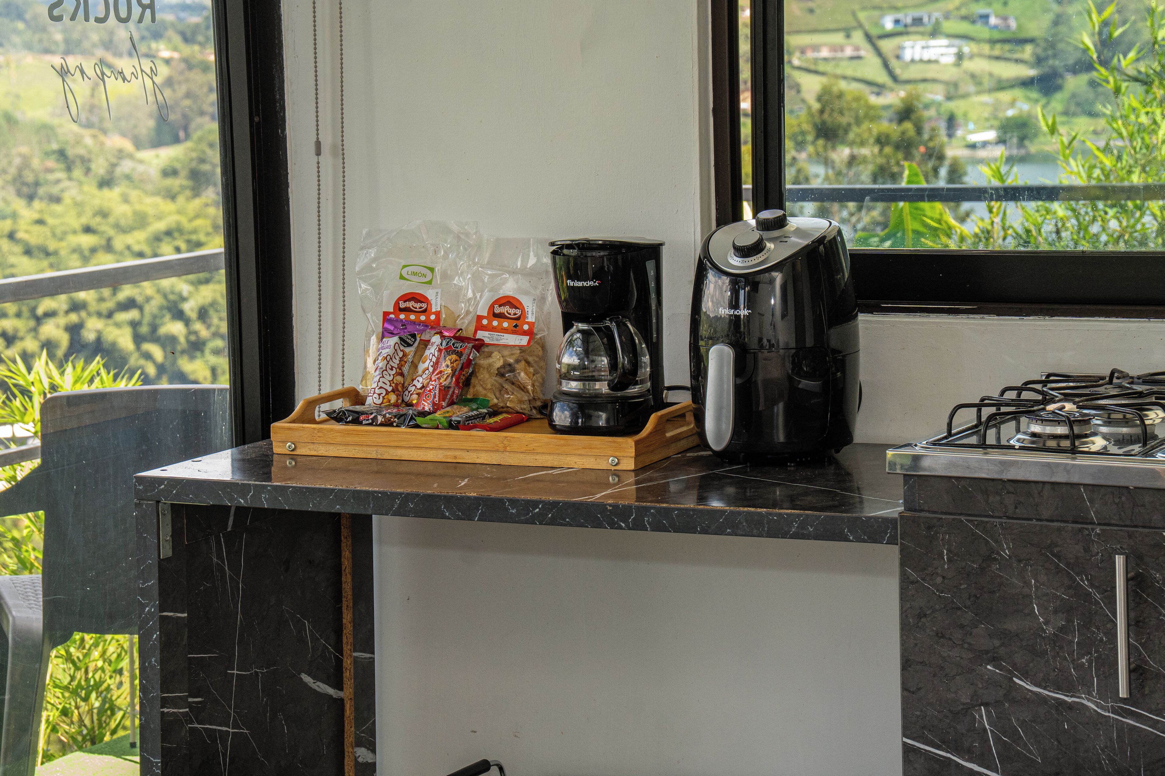 Un taulell de cuina amb una cafetera, un plat de tapes i vista a un paisatge verd a través d'una finestra.