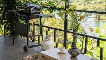 Un balcón con una parrilla, un tanque de gas, una mesa con una taza y vistas a un lago y follaje.