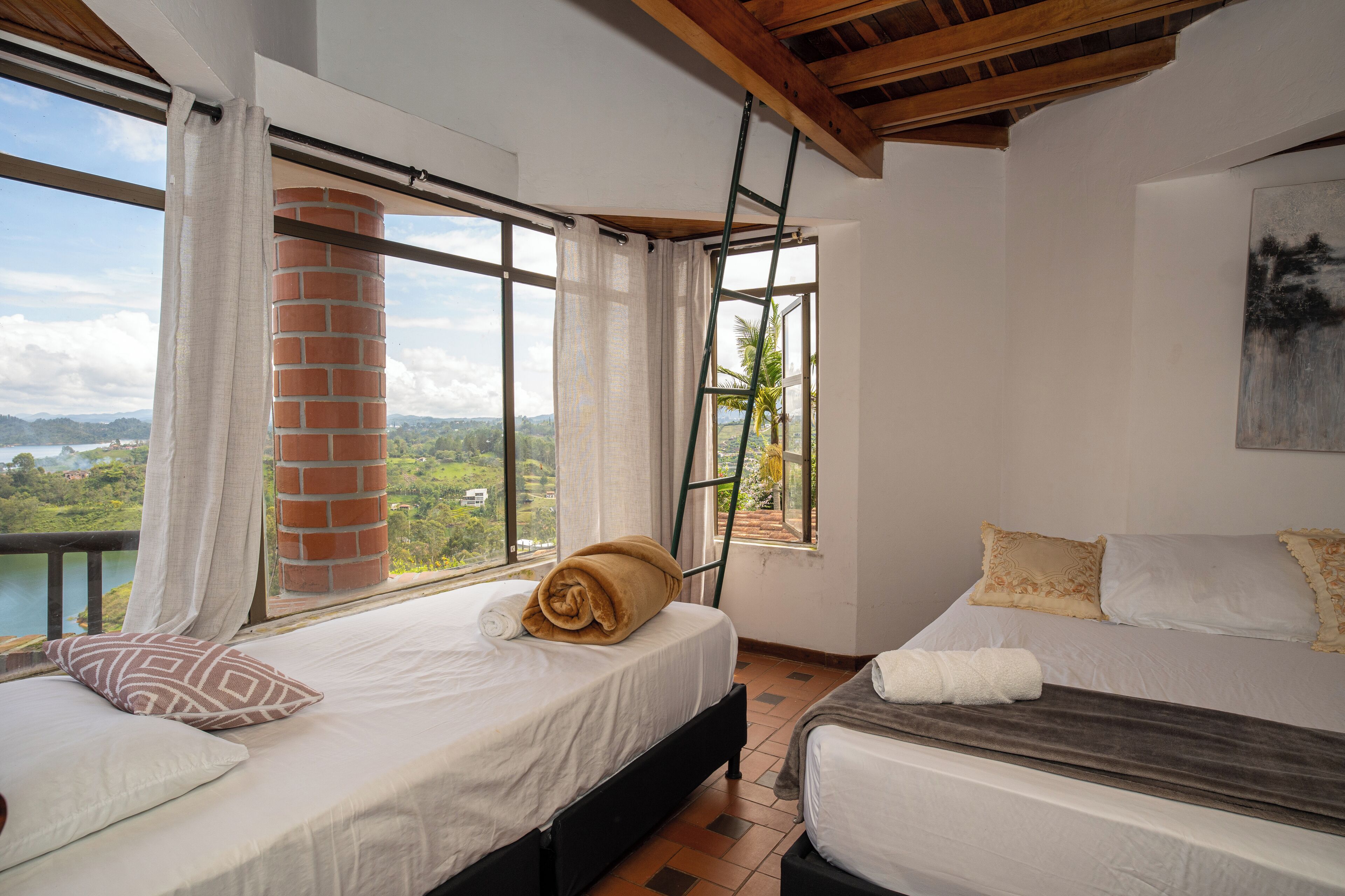A bedroom with two beds, large windows, a brick chimney, and a view of the outdoors.