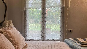 A bedroom with a bed, pillows, a bedside table, and a window with lace curtains. - Il Poggetto di Barbara - Casa Bianca (Scansano)