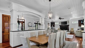 A spacious living area with a wooden dining table, white upholstered chairs, a fireplace, and a television. - Award-Winning Resort Amenities | Fishers Alley 2 by Wild Dunes (Isle Of Palms)