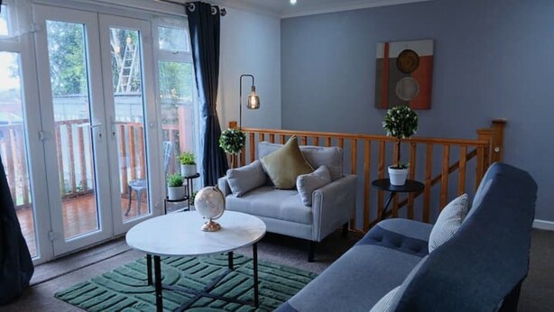 Una sala de estar con un sofá gris, una mesa redonda y un sillón azul. Se ve una escalera con barandilla de madera que lleva a un balcón.