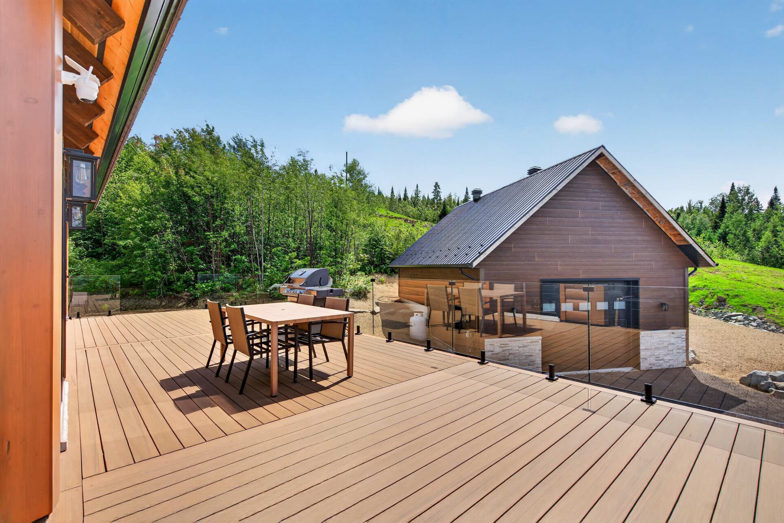 Une terrasse en bois avec une table et des chaises, un barbecue et un espace agrémenté d’une cheminée fermée par des parois de verre.