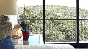 Un salón con un sofá color beige, una almohada azul, un jarrón y una planta en maceta. Un ventanal ofrece vistas a un paisaje montañoso.