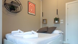 Ein ordentlich bezogenes Bett mit weißen Leinen und braunen Kissen, eine Wandpflanze, ein gerahmtes Kunstwerk und ein Nachttisch mit einer Pflanze.