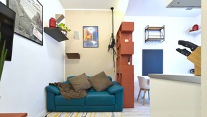 Ein kompakter Wohnbereich mit einem petrolfarbenen Sofa, einem gemusterten Teppich und einer Küchenzeile mit Spüle und Geschirr.