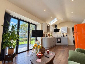A modern kitchen with a flat-screen TV, a dining area with a table and chairs, and a large window with a view of greenery. - Luxury Rustic Barn Retreat in Llanrwst (Llanrwst)