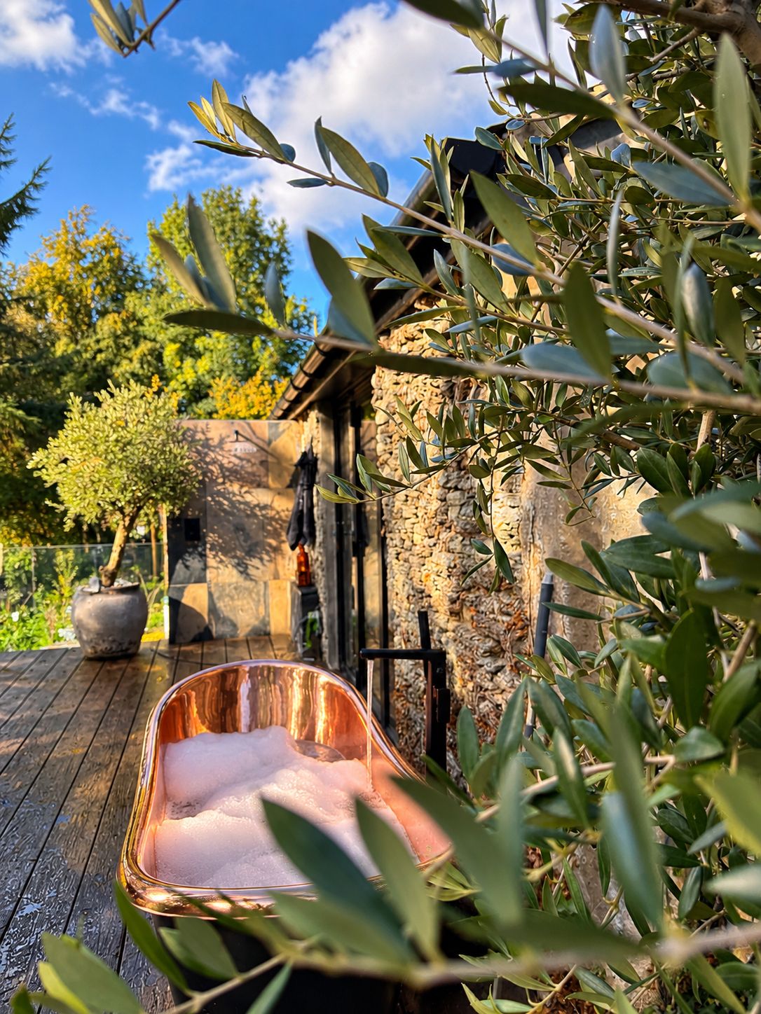 Un entorno rústico al aire libre con un muro de piedra, una terraza de madera y una pila de cobre llena de agua.