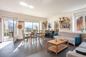 A spacious living area with a dining table, sofa, and bookshelf, large windows, and a piano.
