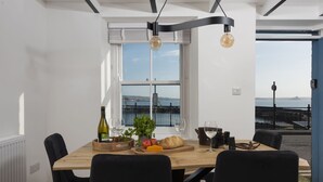 A dining area with a wooden table, black chairs, a bottle of wine, glasses, and a view of the sea through large windows. - Harbour Watch Cottage – Sea View Cottage in Newlyn, Cornwall (Newlyn)