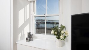 A window with a view of a dock and water, a white vase with white flowers, and a flat-screen TV on a white shelf. - Harbour Watch Cottage – Sea View Cottage in Newlyn, Cornwall (Newlyn)