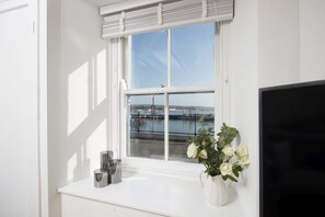 Una ventana con vista a un muelle y al agua, un jarrón blanco con flores blancas, y un televisor de pantalla plana sobre un estante blanco.