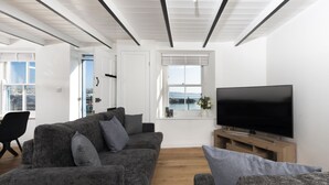 A modern living room with a grey sofa set, a flat-screen TV, and a window with a view of the sea. - Harbour Watch Cottage – Sea View Cottage in Newlyn, Cornwall (Newlyn)