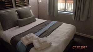 A neatly made bed with a striped bedspread, pillows, and a folded towel. There are two framed pictures on the wall, a floor lamp, and a window with curtains.