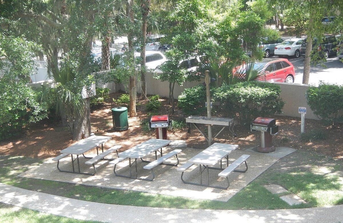 A picnic area with tables and grills, surrounded by trees and a parking lot.