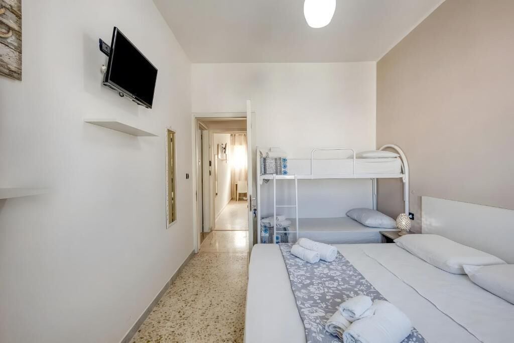 A hotel room with a bunk bed, a flat bed, a television mounted on the wall, and a hallway leading to another room.