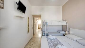 A hotel room with a bunk bed, a flat bed, a television mounted on the wall, and a hallway leading to another room.