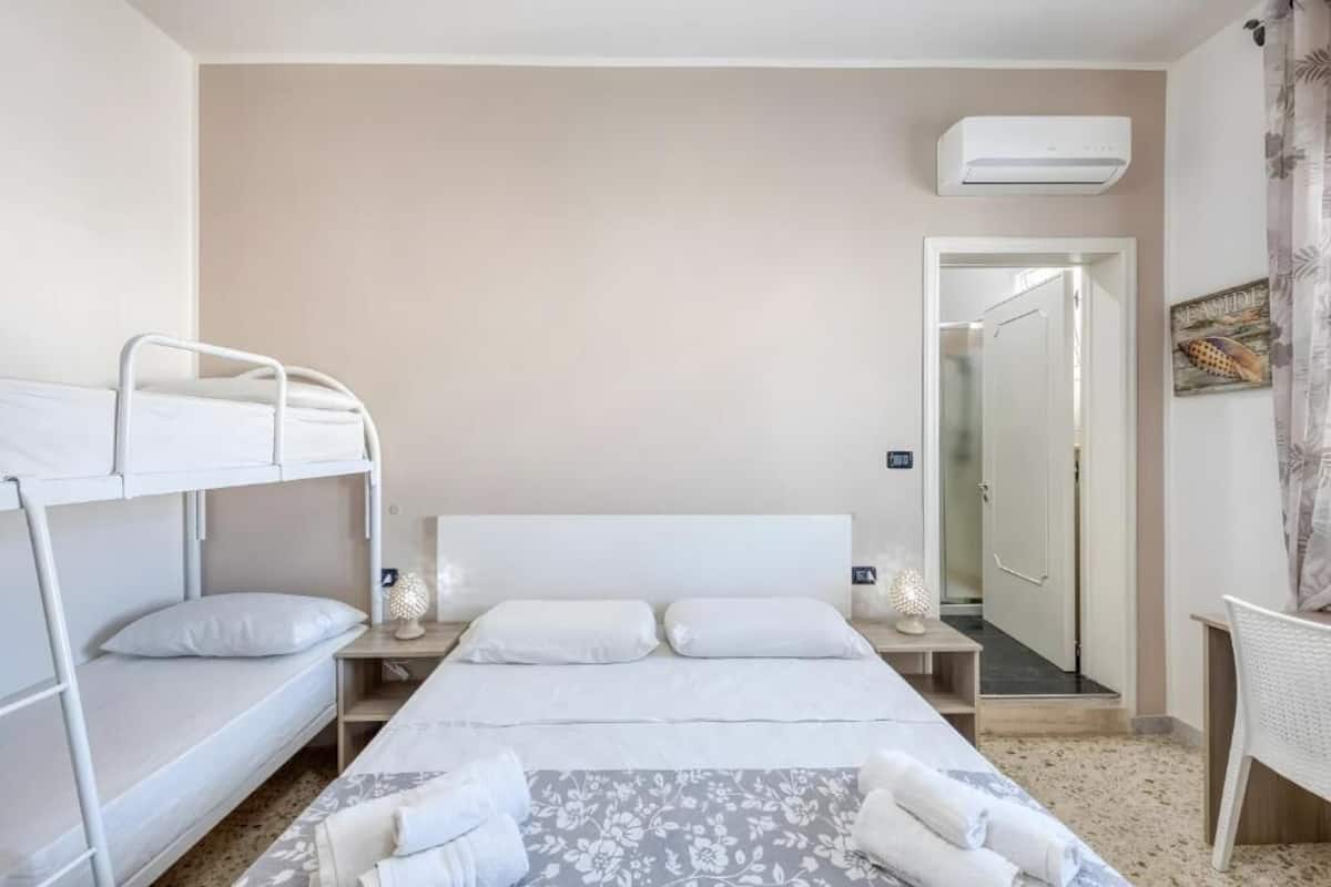 A hotel room with a bunk bed, a single bed, a bedside table with a lamp, a bathroom door, and a shower area.