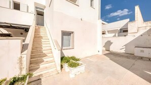A white building with a staircase leading to a door, a small window, and a planter with greenery. - Il Comandante (Torre San Giovanni)