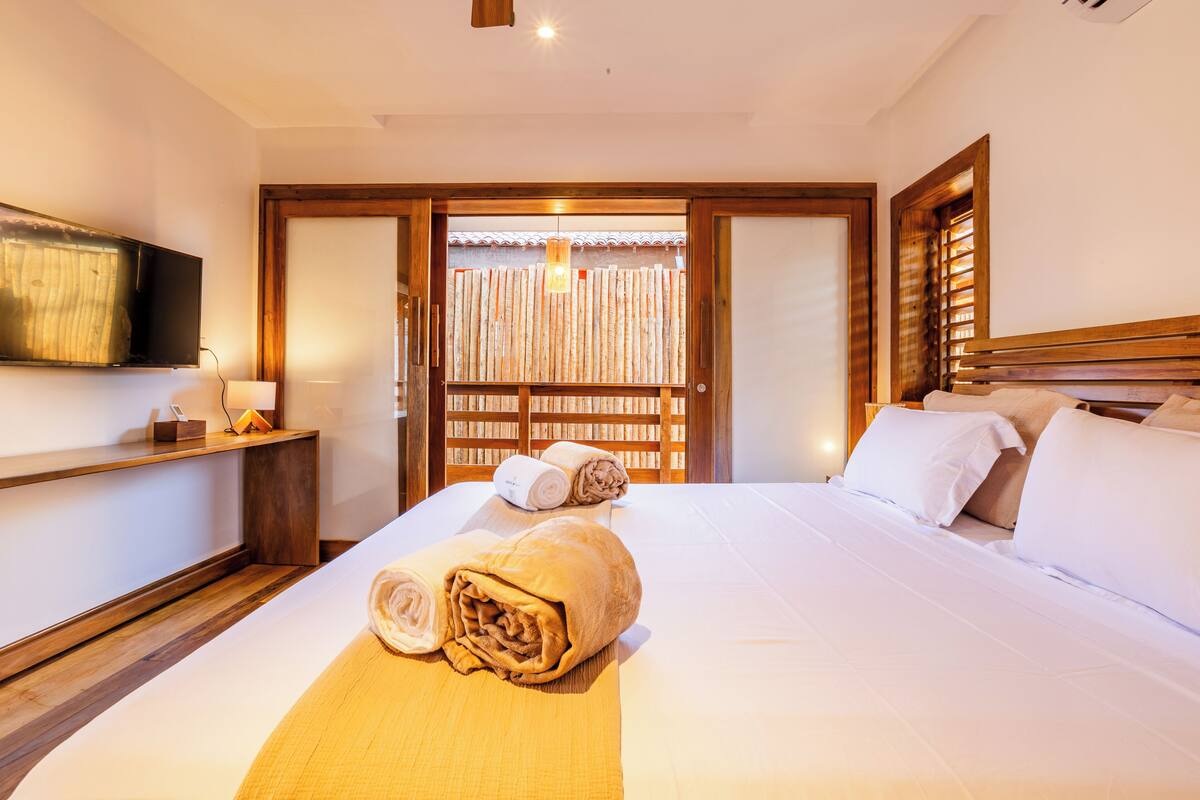 A hotel room with a neatly made bed, a television on the wall, and a wooden door with a view of a bamboo fence.