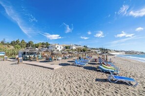 Uma praia com espreguiçadeiras, guarda-sóis e um edifício com área de piscina.