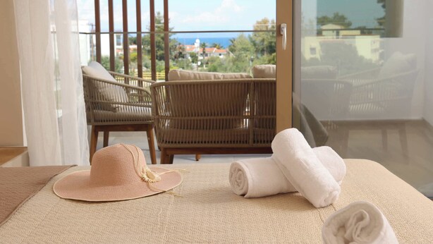 Una habitación con vista al océano, una silla de mimbre, una toalla enrollada y un sombrero de paja.