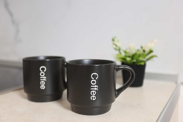 Dos tazas de café negras con la palabra 'Coffee', sobre una encimera con una planta en maceta al fondo.