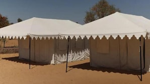 A row of white tents with a scalloped edge, set on sandy ground with trees in the background.
