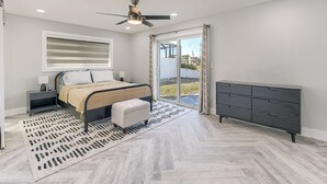 Un dormitorio con cama, mesita de noche, lámpara, ventilador de techo, cómoda y vistas al exterior a través de una puerta de cristal.