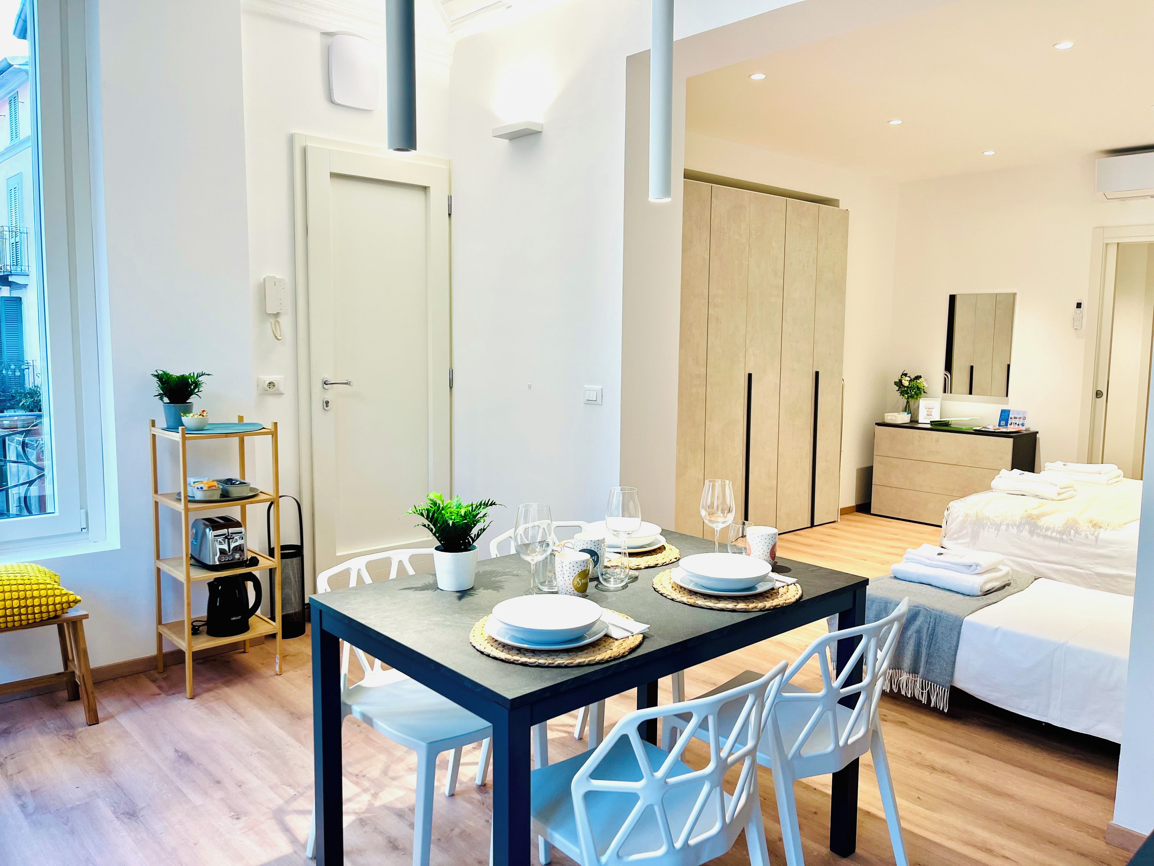 A modern, well-lit room with a dining table set for two, a bed with white bedding, and a wooden wardrobe.
