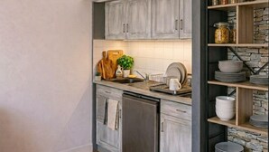 Cucina con mobili grigi, lavastoviglie, tagliere di legno, una pianta e uno scaffale in pietra con barattoli.