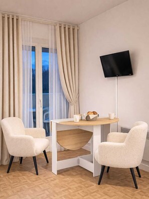 A room with a TV mounted on the wall, a round table with chairs, and a window with curtains. - Studio in Barbotan-les-Thermes - Balcony, Parking & 300 m from the Spa (Cazaubon)