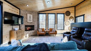 A cozy room with a fireplace, leather seating, a flat-screen TV, and large windows. - New! Private Heated Pool, Hot Tub, Game Room, Fire Pit & Bunk Room - Near Trails (Broken Bow)