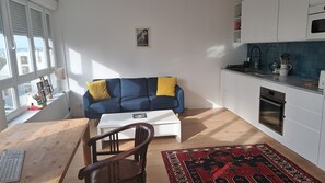 A compact living space with a sofa, a small table, a chair, a kitchen area, and a view through large windows. - APARTMENT 50m2 MOUZAÏA/PLACE DES FÊTES (Paris)