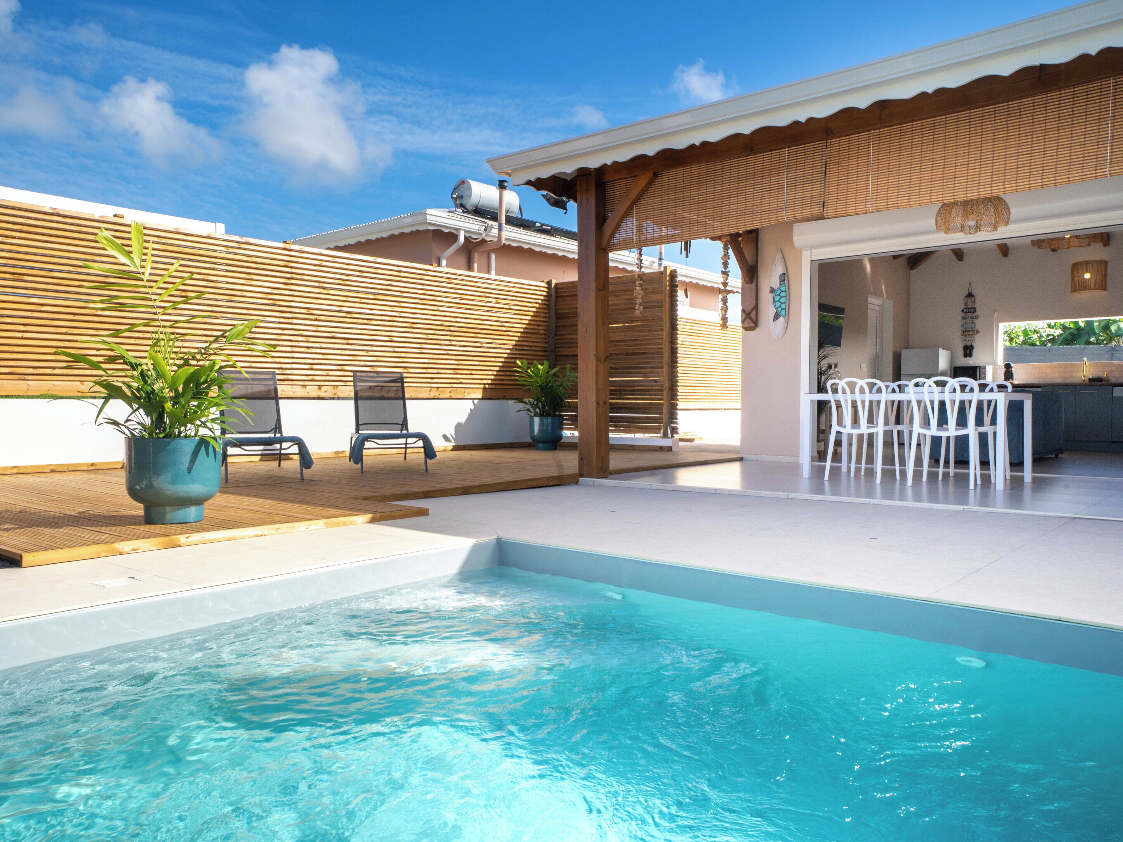 A swimming pool with a wooden deck, a covered outdoor seating area, and a kitchen visible through an open door.