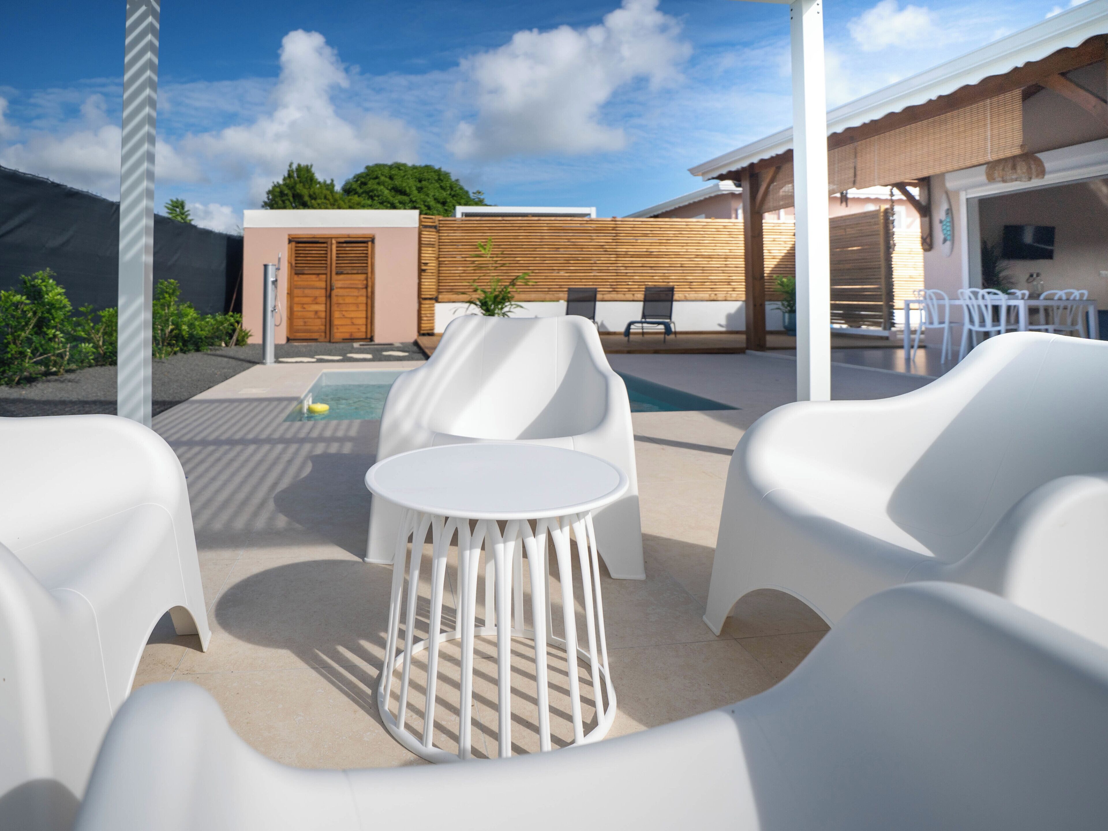 Un espace extérieur moderne aménagé avec des chaises blanches et une table, une piscine, et un bâtiment en bois en arrière-plan.