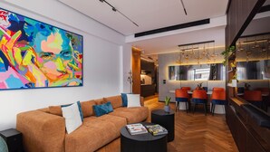 Een moderne woonkamer met een bruine bank, een salontafel en een kleurrijk abstract schilderij. Er is een eethoek met oranje stoelen en een keuken op de achtergrond.
