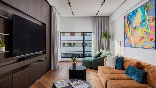 Een moderne woonkamer met een flatscreen televisie, een bruine bank, een groene fauteuil, een ronde salontafel met boeken en een groot abstract schilderij aan de muur.