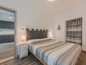Ein Schlafzimmer mit Bett, Nachttischlampen, einem Nachttisch, einem Kleiderschrank und Blick auf eine Küche.