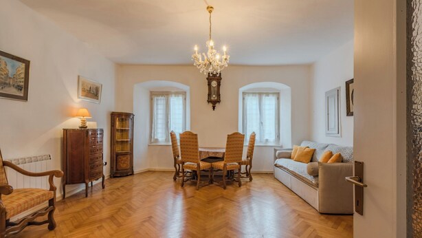 Ein geräumiges Wohnzimmer mit Holzdielenboden, einer Kronleuchter, einem Esstisch mit Stühlen, einem Sofa und einem Schrank.