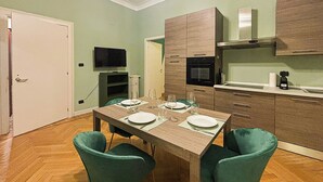 A dining area with a wooden table, four teal chairs, and a kitchen with wooden cabinets and a microwave. - Flatsharing - Scarlatti - 2 camere (Milan)