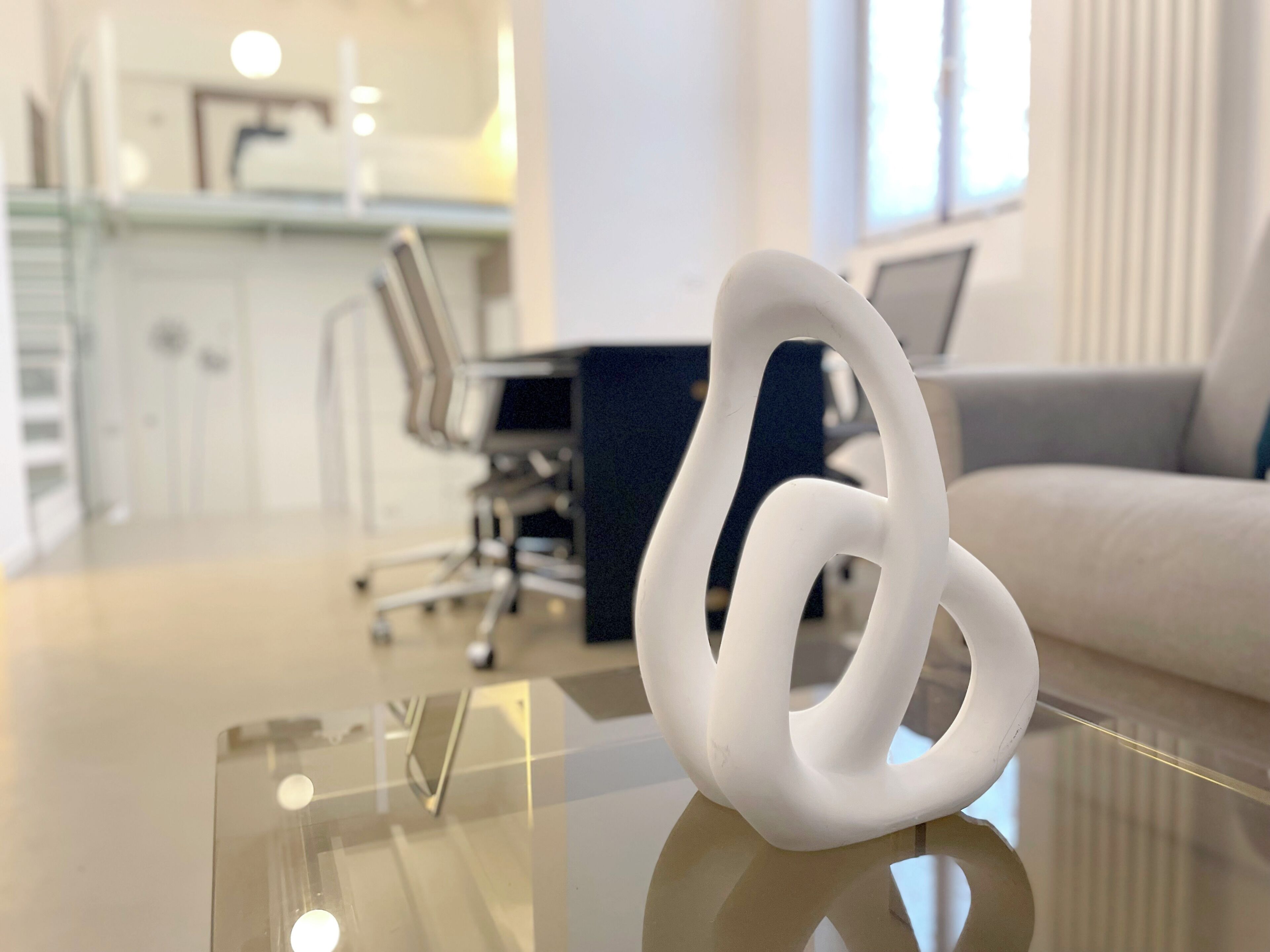 Ein modernes Interieur mit einem Glastisch, einer weißen abstrakten Skulptur, einem weißen Sofa und einer Küchenzeile im Hintergrund.
