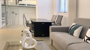 Ett modernt vardagsrum med ett soffbord i glas, en grå soffa med en dekorativ kudde och ett svart skrivbord med stolar.