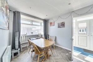 A modern dining room with a wooden table and chairs, large windows with curtains, and a door with a mat. - 3 Bedrooms Sleeps 6 Spacious Chalet Log Burner (Bridlington)